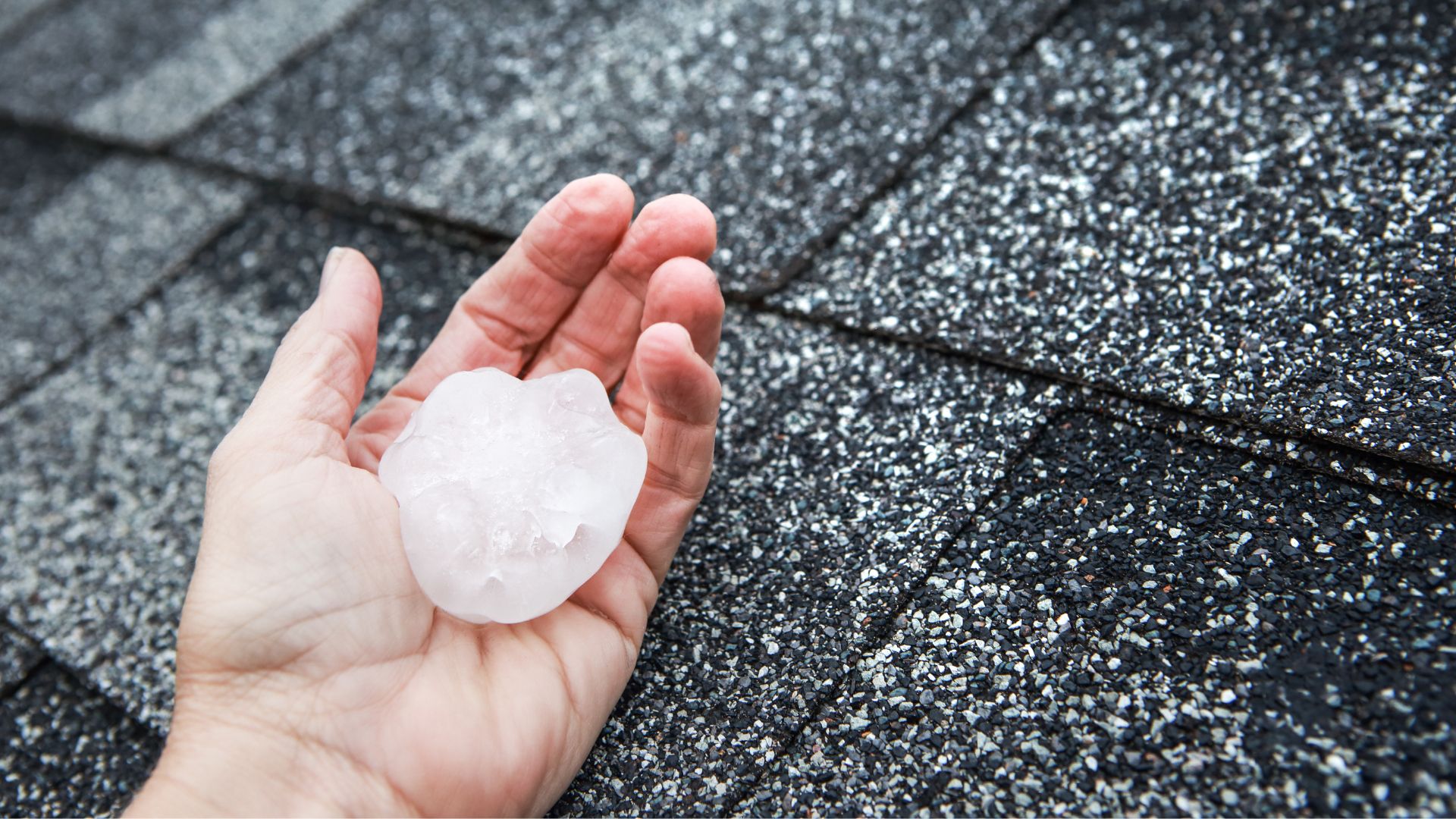 A hand is holding a large piece of hail with a roof in the background.