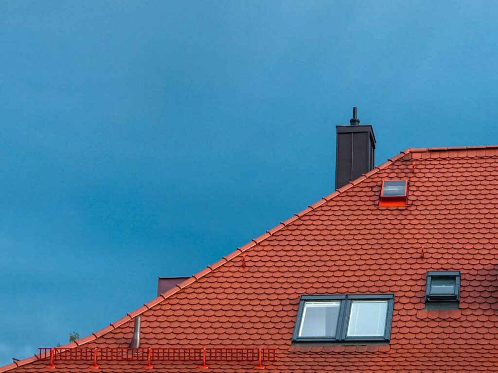 A red-tiled roof with two skylights and a chimney against a clear blue sky.