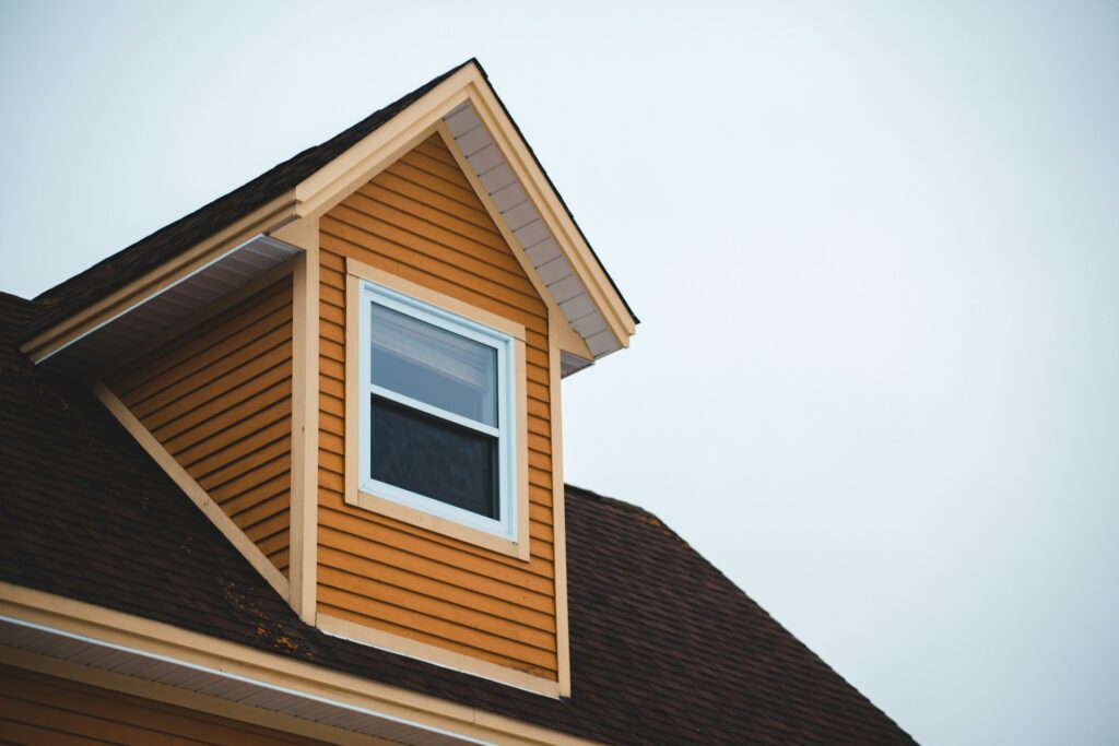 A dormer with a single window on a house with brown siding and a dark brown roof, set against a cloudy sky.