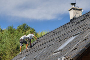 Two workers on a sloped roof install solar panels under a clear sky, with trees in the background.