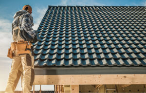 A construction worker in a plaid shirt and tool belt examines a newly installed dark tiled roof on a sunny day.