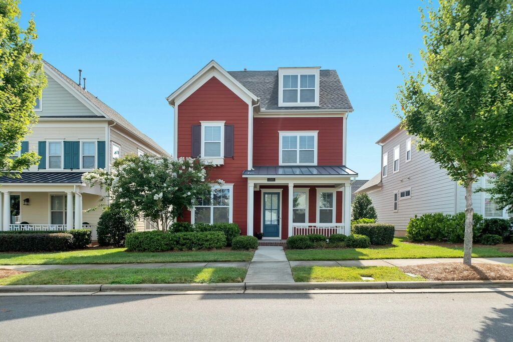 Red two-story house with white trim and a porch, surrounded by greenery. Flanked by similar houses. Clear blue sky in the background. Street view.
