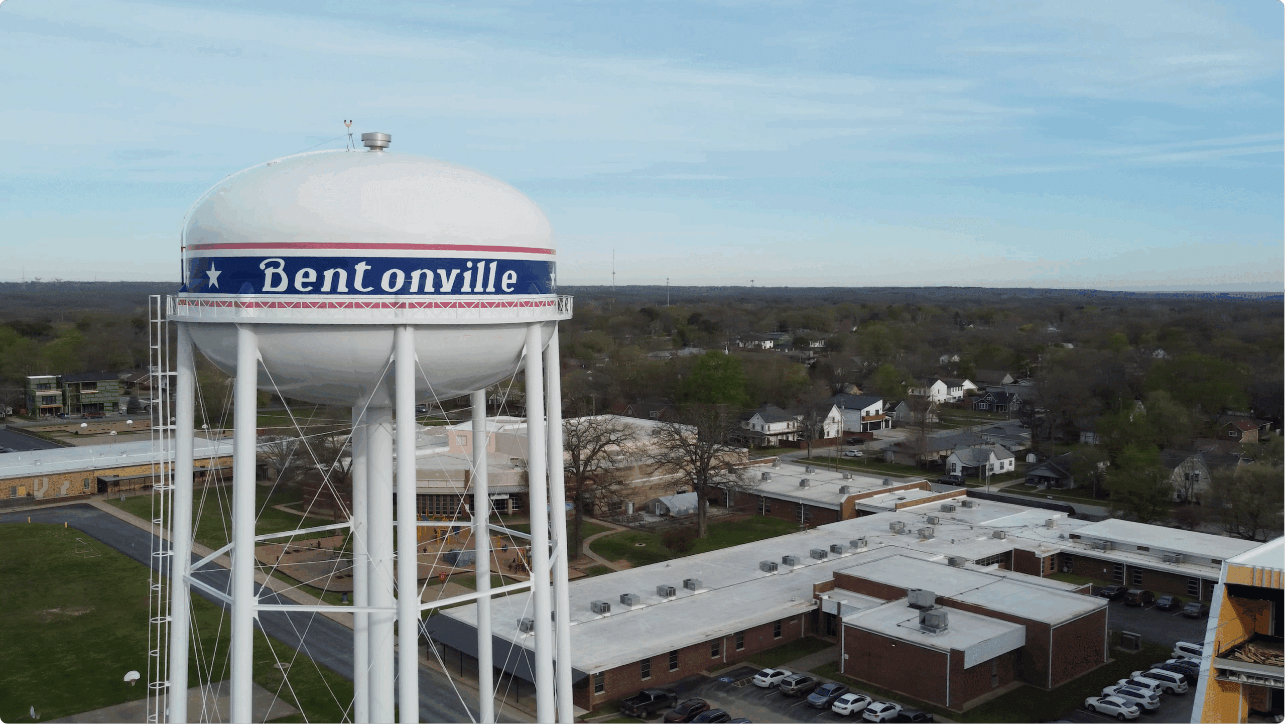 A white water tower labeled "Bentonville" stands near a school building and residential neighborhood under a cloudy sky.