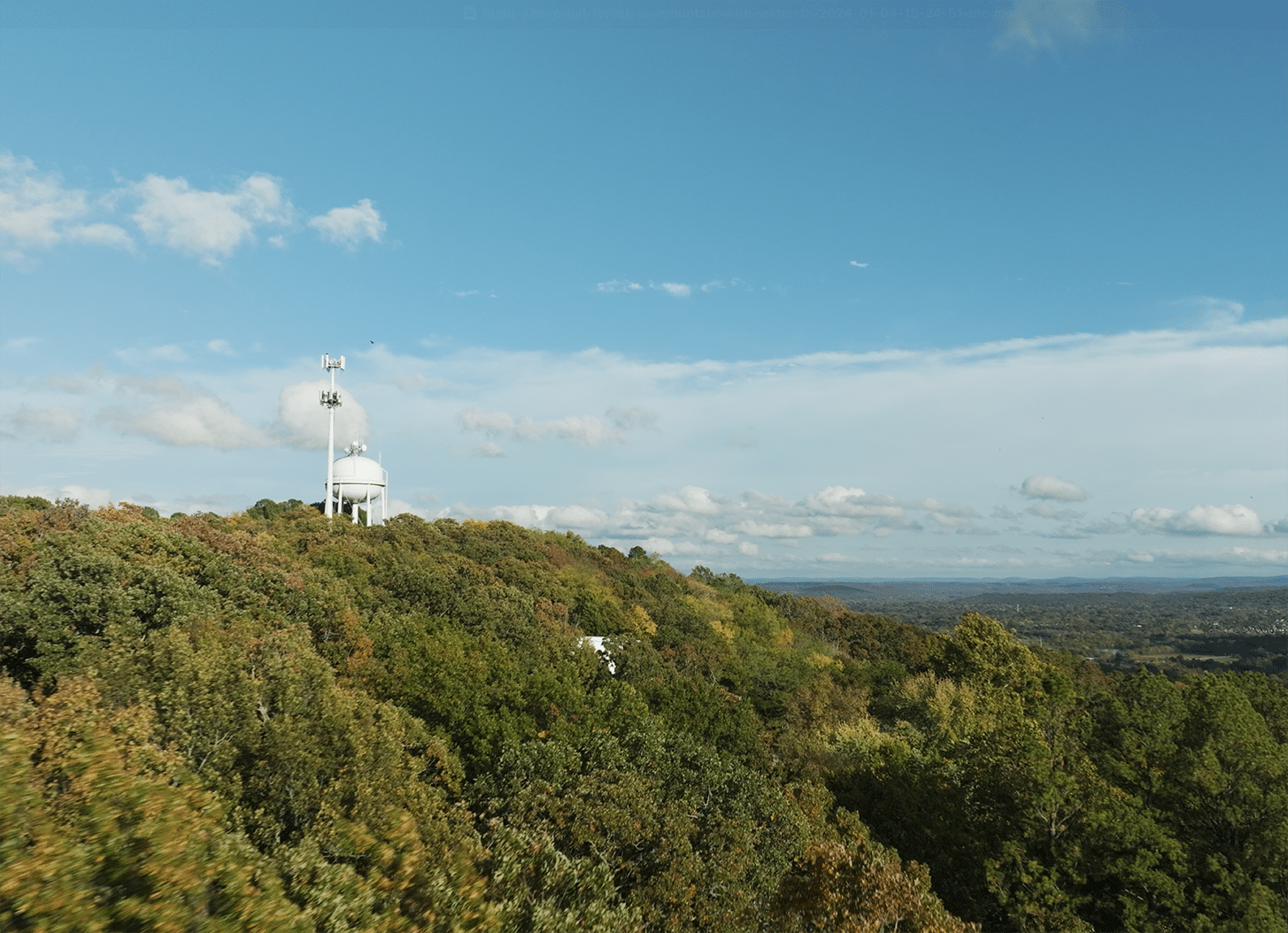A white observatory dome stands on a wooded hilltop under a blue sky with scattered clouds, overlooking a distant landscape.