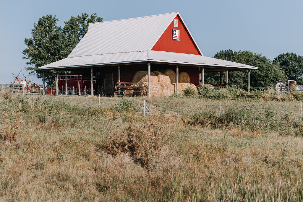 A red barn with a gray roof and hay bales stacked under an overhang sits in a grassy field, with trees and fencing in the background.