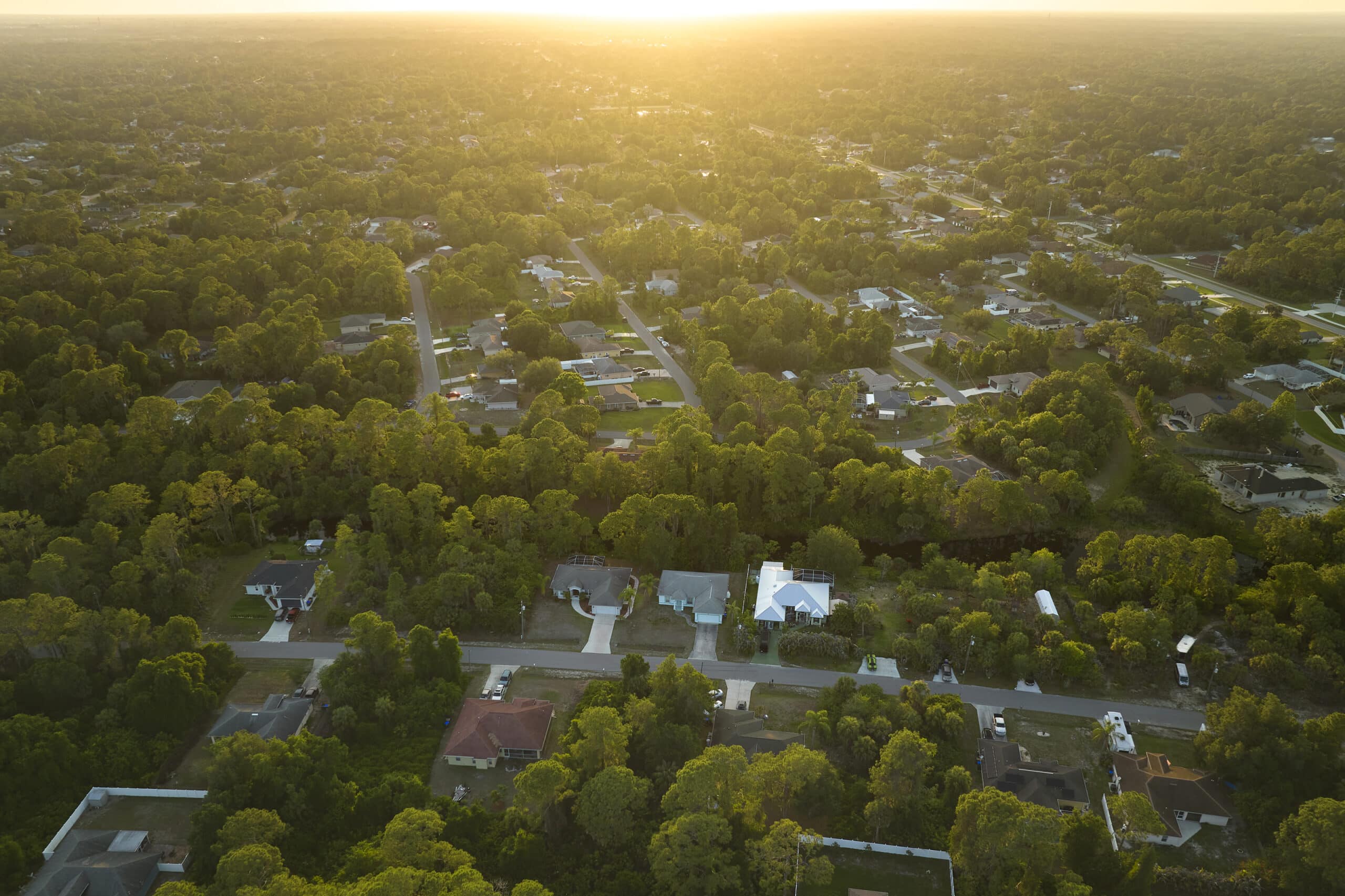 Aerial view of a suburban neighborhood with houses, trees, and roads at sunset. The sunlight creates a warm, golden glow over the landscape.