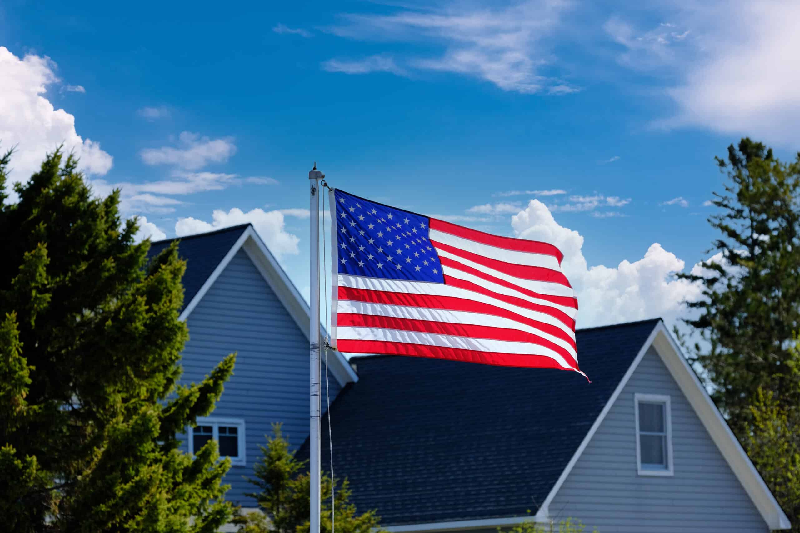 An American flag waves on a flagpole in front of a house with a blue sky and clouds in the background.