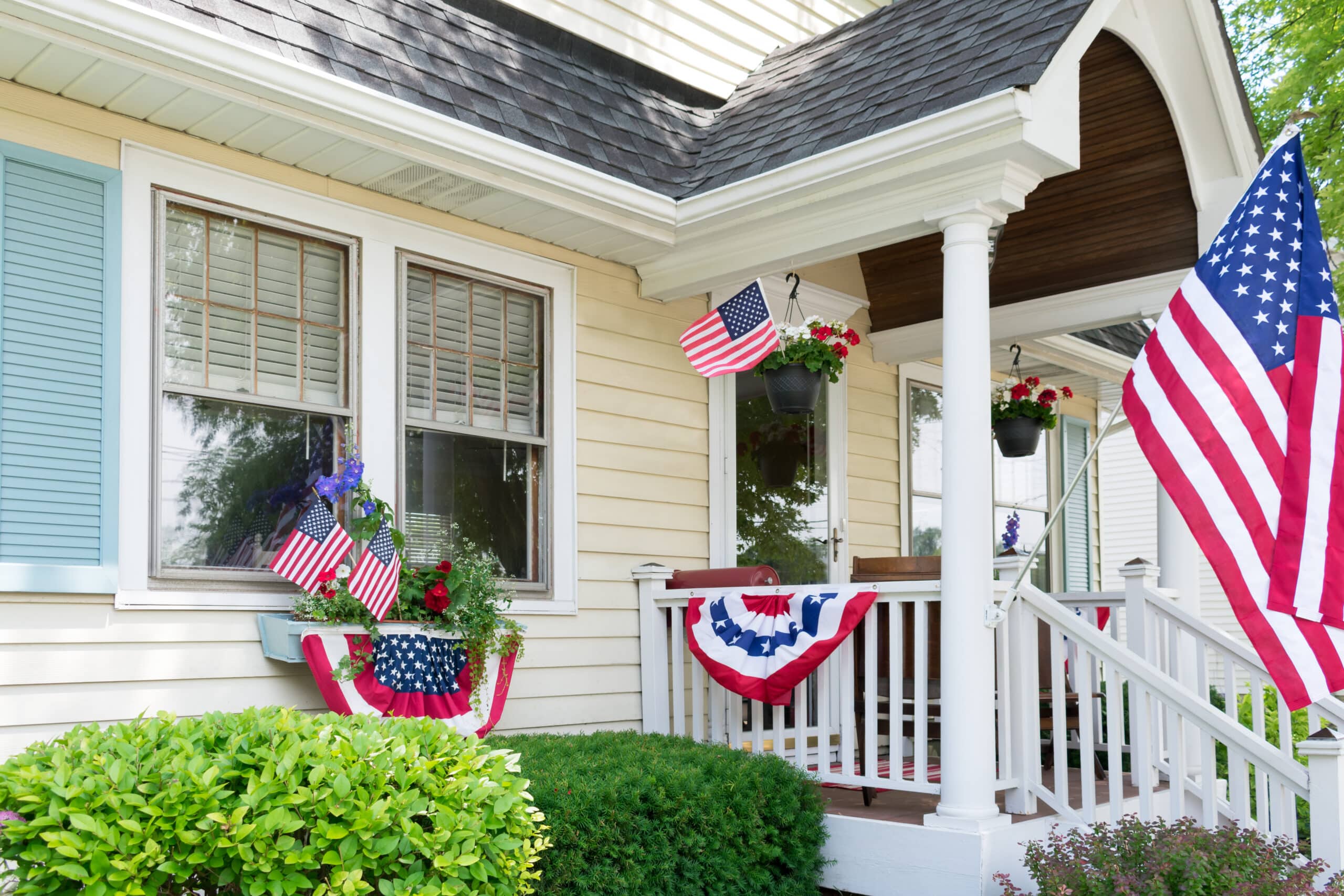 A house decorated with American flags and red, white, and blue bunting on the porch, with flowers and greenery in front.