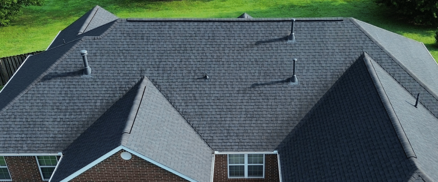 Aerial view of a house with a new dark gray asphalt shingled roof, multiple roof peaks, and several vent pipes, surrounded by green grass. A project completed by Davids Roofing.