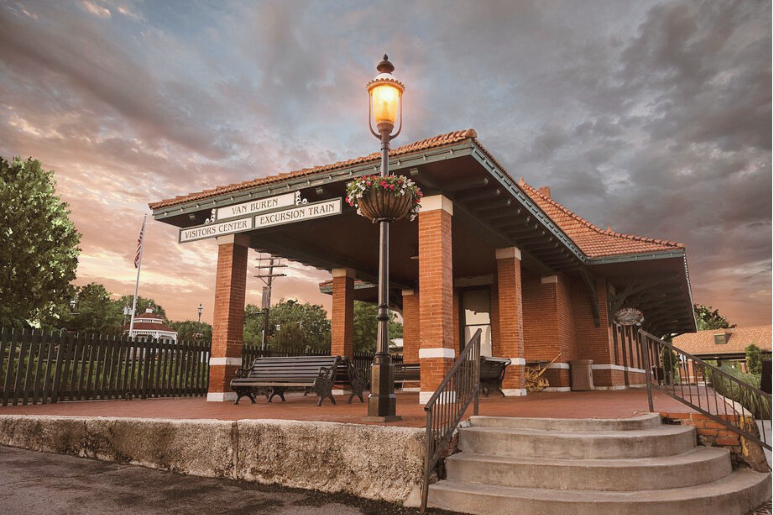Van Buren Arkansas tourist center. A brick train station with a covered platform, benches, hanging flower baskets, and a lit streetlamp, set against a cloudy sky at sunset.