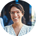 Woman with short dark hair and glasses smiling, wearing a white blouse with dark dots, standing outdoors in a blurred urban setting.