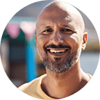 A man with a shaved head and beard, wearing a light yellow shirt, smiles outdoors with a blurred background.