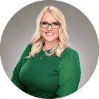 A woman with long blonde hair and glasses wearing a green top, smiling at the camera against a gray background.